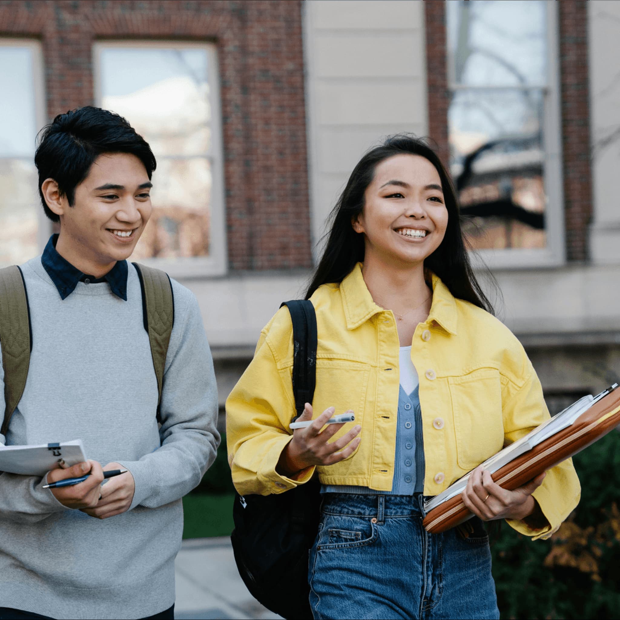 Two students walking outdoors, one in a yellow jacket and the other in a gray sweater, both smiling and carrying notebooks.