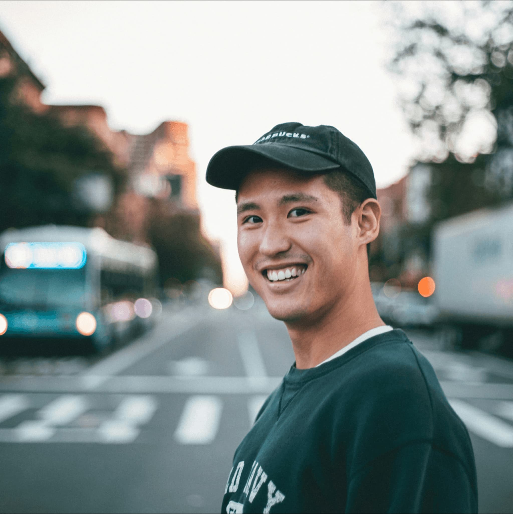 Smiling young man wearing a cap, standing on a city street with blurred vehicles in the background at sunset.