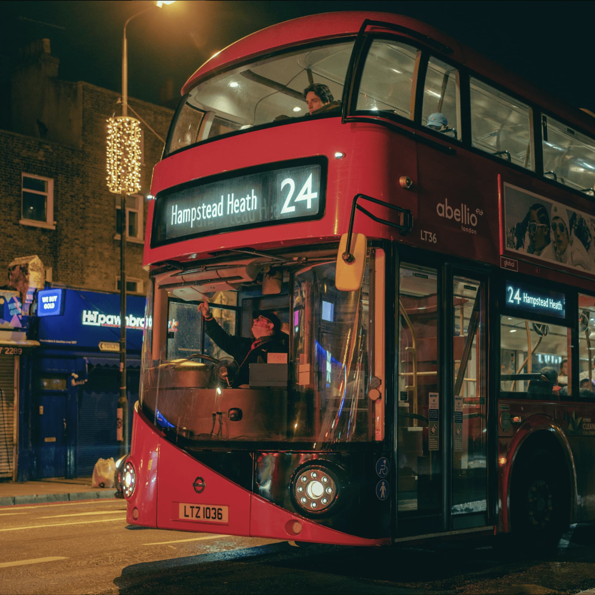 London bus on route 24, labeled "Hampstead Heath," featuring a driver and illuminated city street surroundings at night.