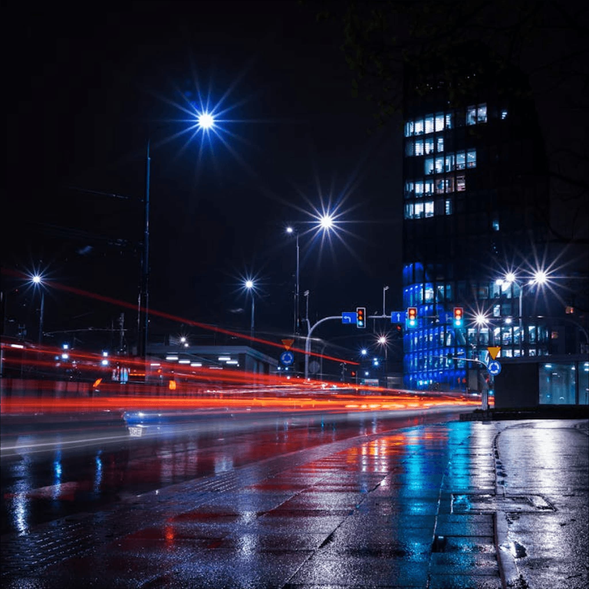 City street at night with illuminated buildings and blurred car lights reflecting on a wet pavement.