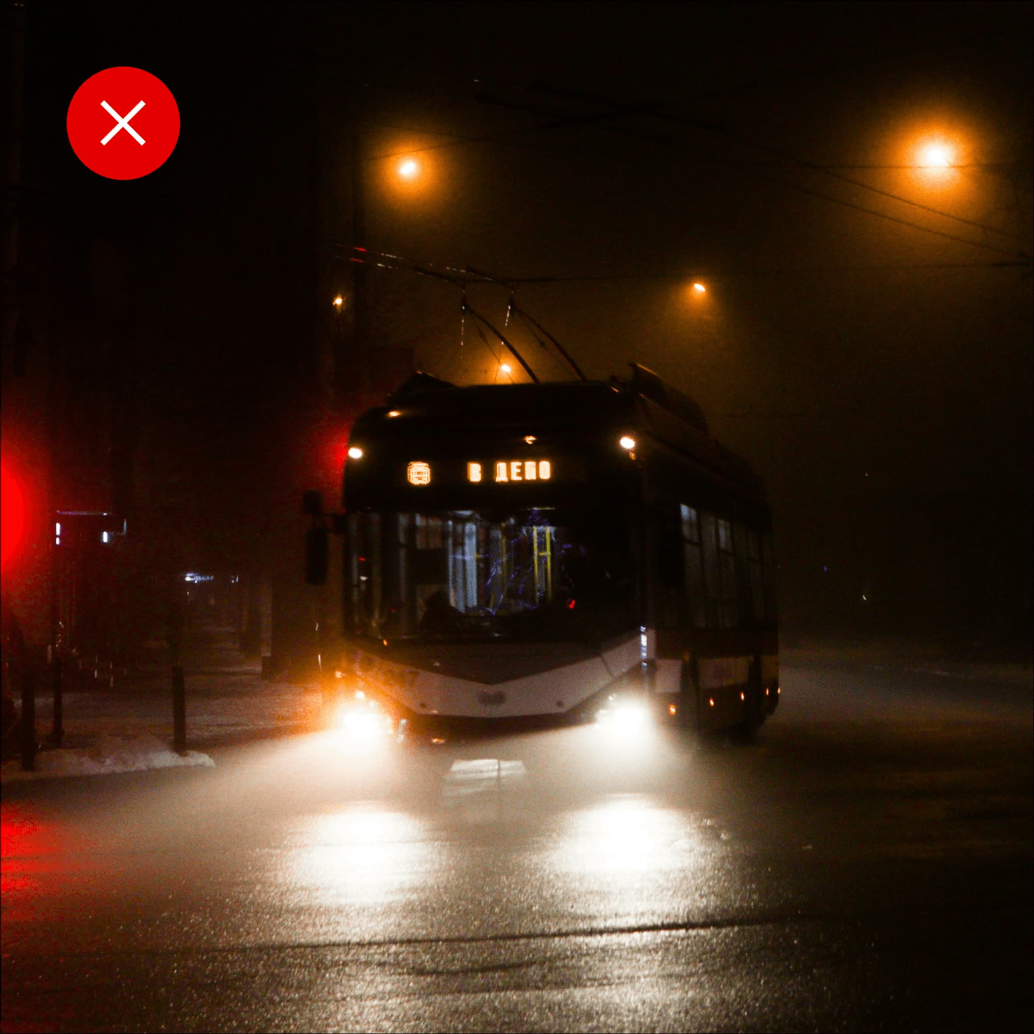 A city tram illuminated by streetlights navigating through a wet, foggy night. Traffic lights visible in the foreground.